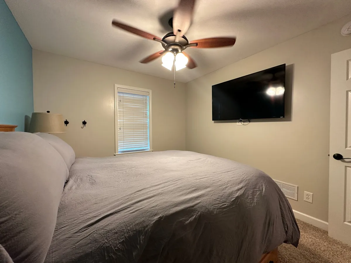 A serene bedroom with a comfortable bed dressed in gray bedding, a ceiling fan above, a wall-mounted TV, and natural light filtering through blinds. The ambiance is peaceful and inviting, ideal for restful nights.
