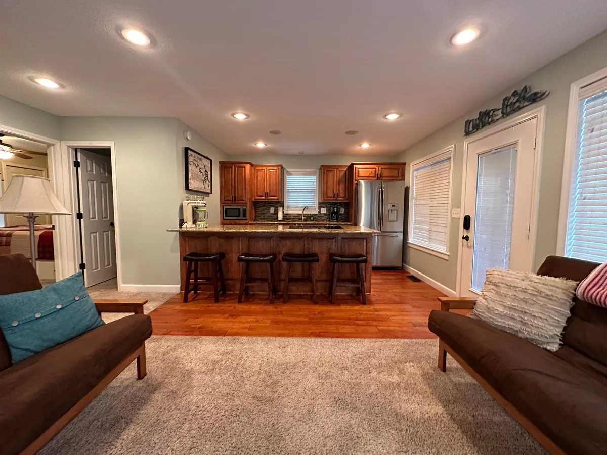 This image highlights a welcoming open-plan living space featuring a kitchen island with bar stools, a cozy sofa with decorative pillows, and warm lighting from a nearby lamp. The layout encourages conversation and togetherness, making it ideal for gatherings, meal prep, and relaxation.