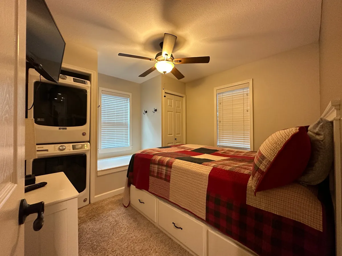 A warmly lit bedroom featuring a bed with a quilt, a cozy window seat ideal for reading or relaxing, a television for unwinding in the evenings, and a stacked washer-dryer tucked into the corner, maximizing hidden storage and organization.