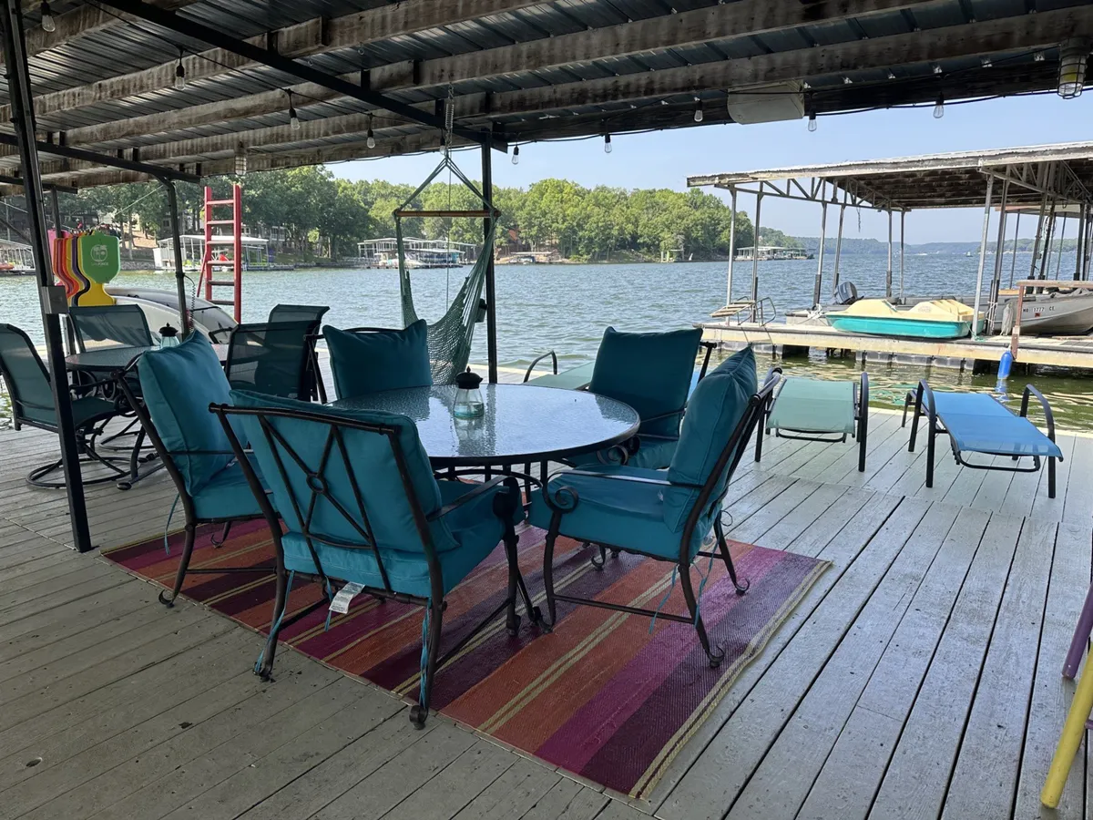 A casual and tranquil exterior deck featuring a round table with chairs, a hammock, a colorful rug, and a picturesque lake view. The setup encourages both relaxation and social gatherings, with areas for dining, lounging, and enjoying the water.