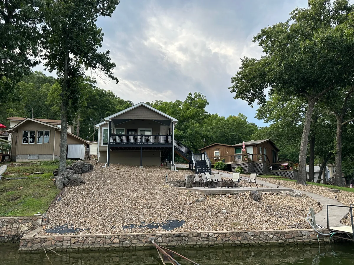 A peaceful lakefront house is shown with an outdoor seating area, rock landscaping, and surrounding trees. The tranquil waters and open sky suggest a serene environment ideal for relaxing, dining, or gathering with friends at sunset.
