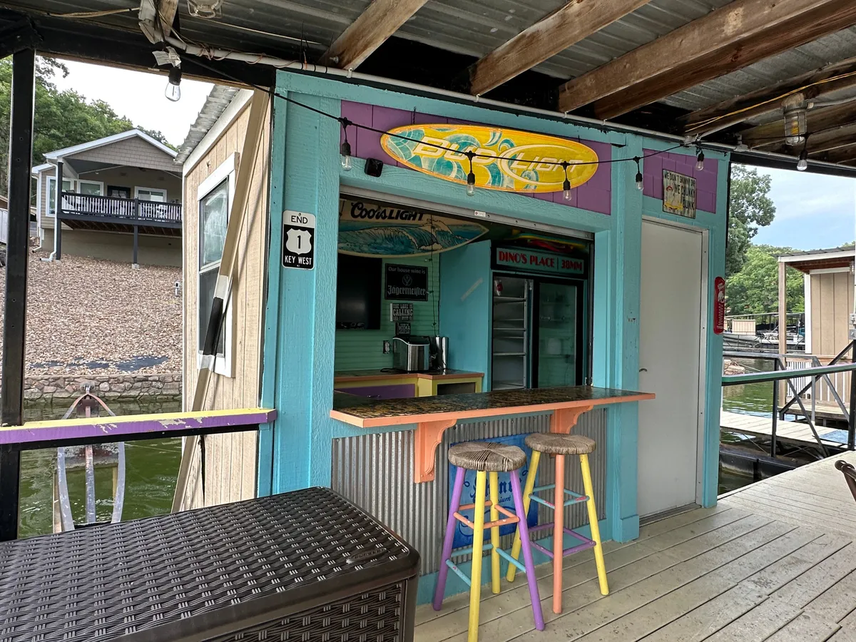 A vibrant, colorful outdoor bar area with surfboard decor, playful stools, and a clear view of the lake. The setup evokes a fun, festive mood and invites socializing in a casual, waterside setting. The lively atmosphere hints at the entertainment available at Bear Bottom Resort.