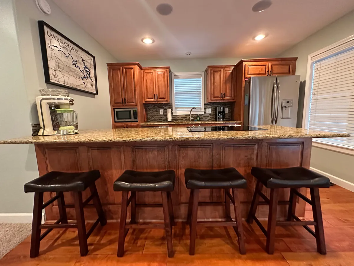 A view of the kitchen with a granite countertop, wooden cabinetry, bar stools, and modern appliances. The setup is ideal for meal preparation, casual dining, and family activities, emphasizing the fully stocked aspect and functionality of the kitchen.