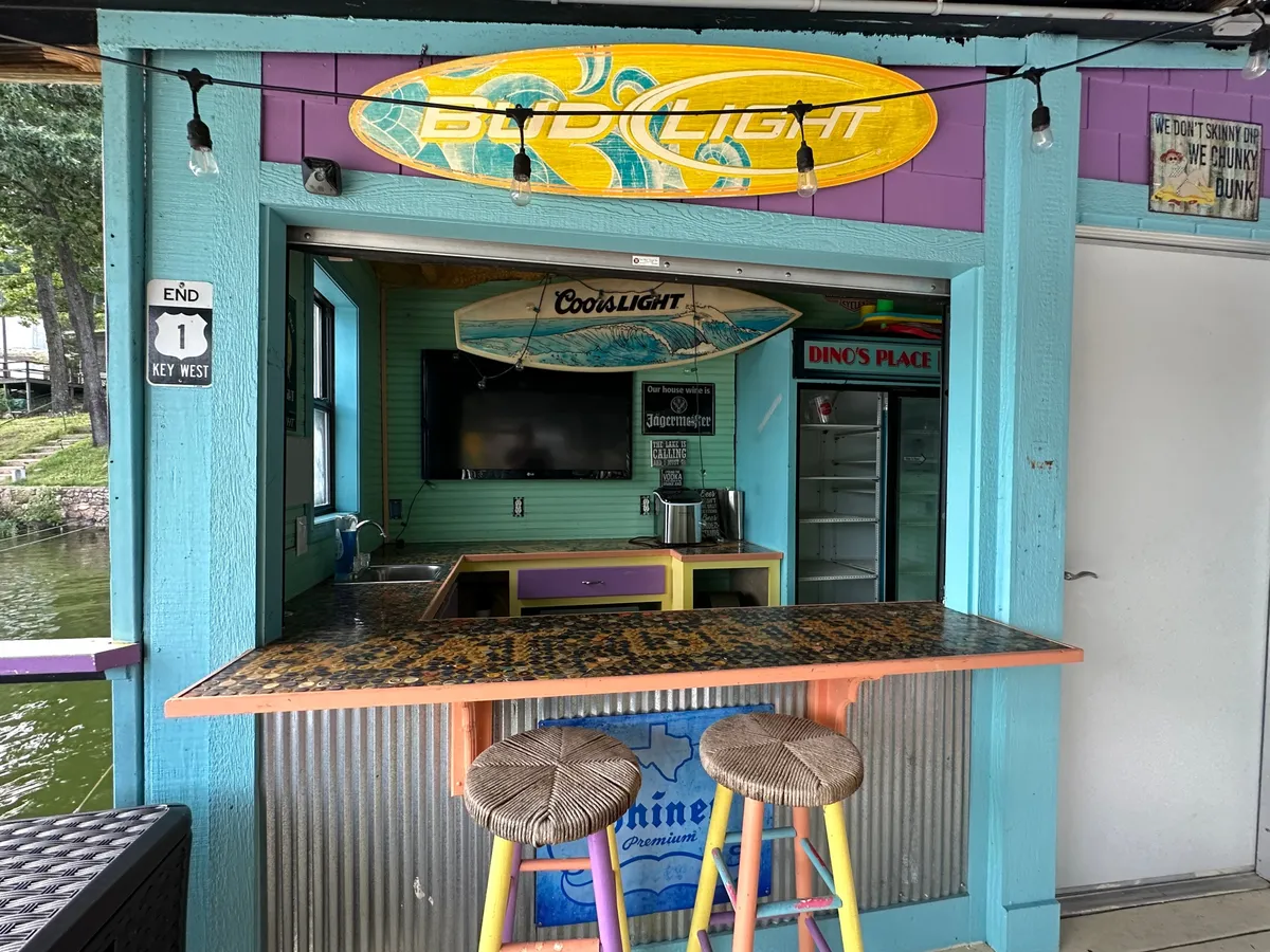A vibrant interior space featuring a bar countertop with seating stools, colorful surfboard decor, and lively walls. The area is well-lit and designed for socializing, with a visible door that could plausibly be equipped with smart security features like a video/audio doorbell and smart locks.