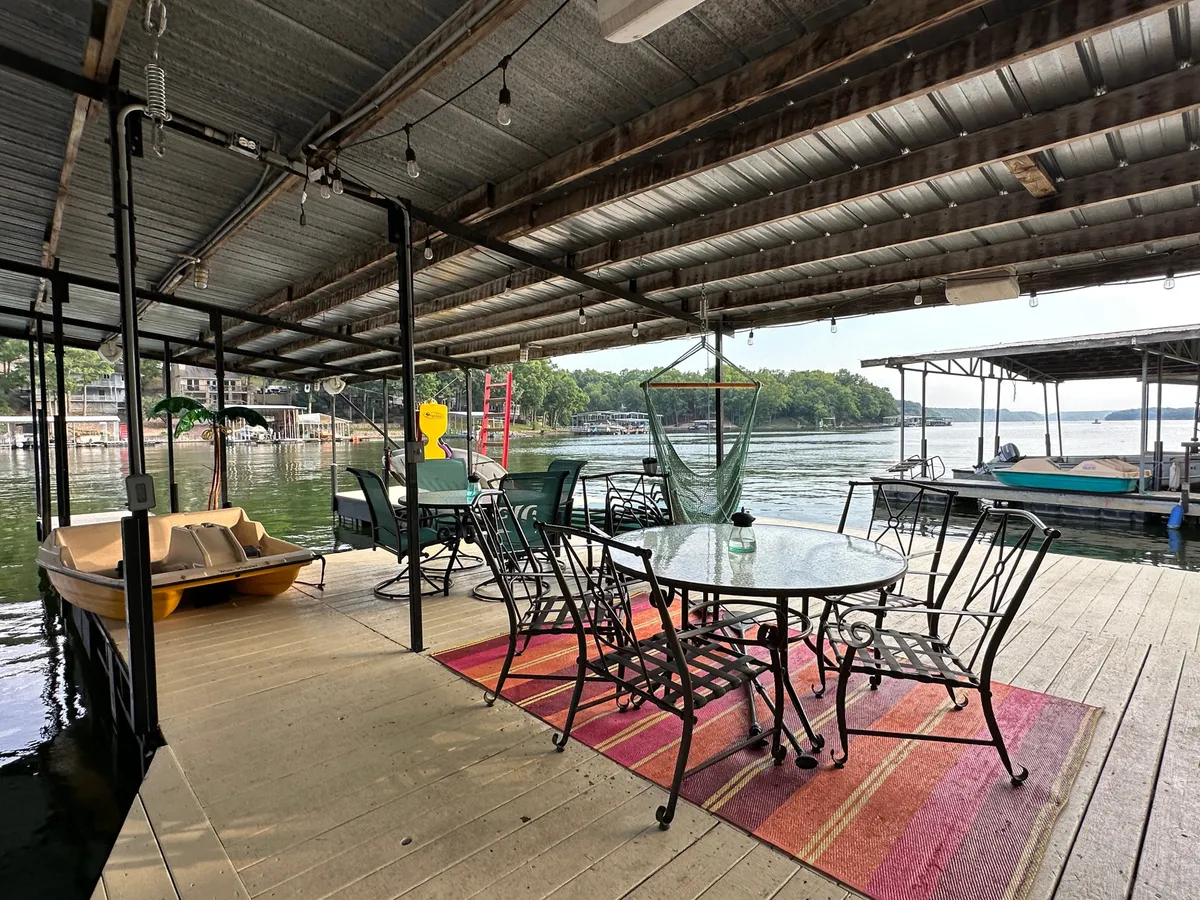 A vibrant outdoor scene with a glass table, metal chairs, a hammock, and boats visible in the water. The area is set up for casual dining, socializing, and relaxing by the lake, with lively touches that suggest both adventure and comfort.